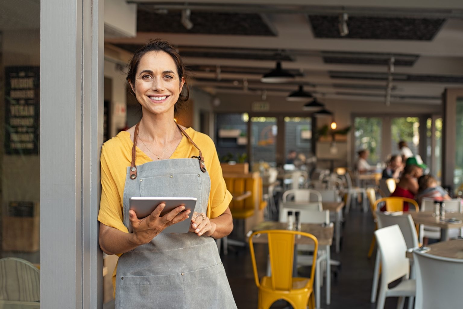Small restaurant owner in apron smiling in front of busy dining room